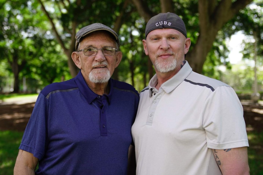 An elderly man wearing a blue t-shirt and hat with a younger man in a white t-shirt