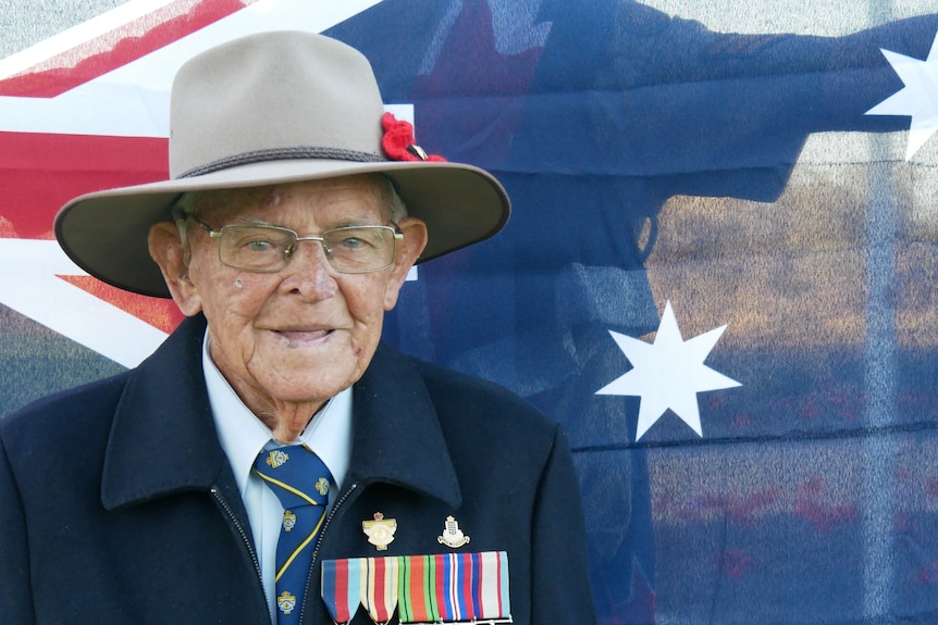 A World War II veteran wears a hat and his badges in front of the Australian flag. 