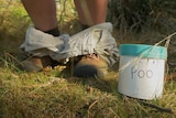 The feet of someone pretending to be going to the toilet in the bush, with a poo pot.