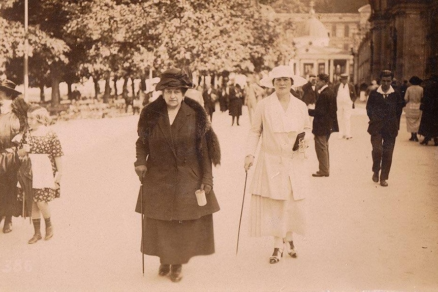 An old photograph of two well-dressed woman, one in black and another in white, pose with canes near a park and crowd.
