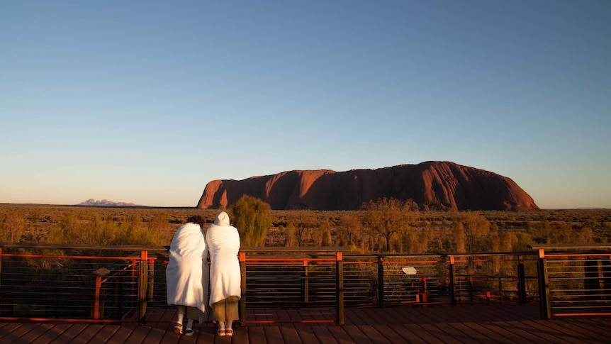 Two people standing in bathrobes look at Uluru at sunrise