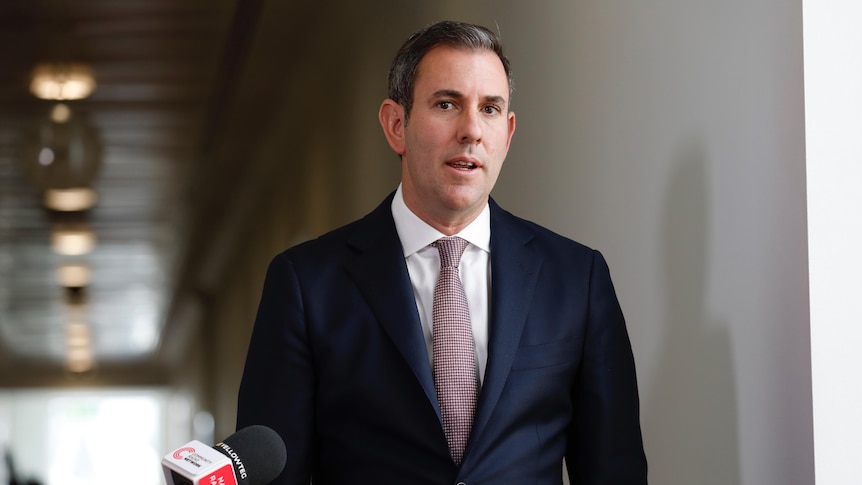 A middle-aged white man in a dark blue suit speaks to reporters in a hallway.