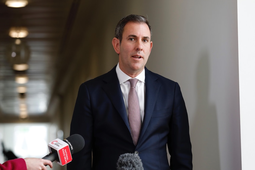 A middle-aged white man in a dark blue suit speaks to reporters in a hallway.