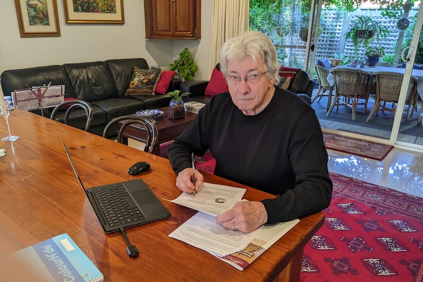 A man with white hair and a black jacket sits at a desk in a house with paperwork around him.