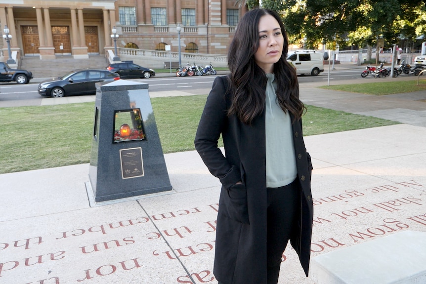 A woman with long, dark hair, wearing a trench coat, standing at a war memorial
