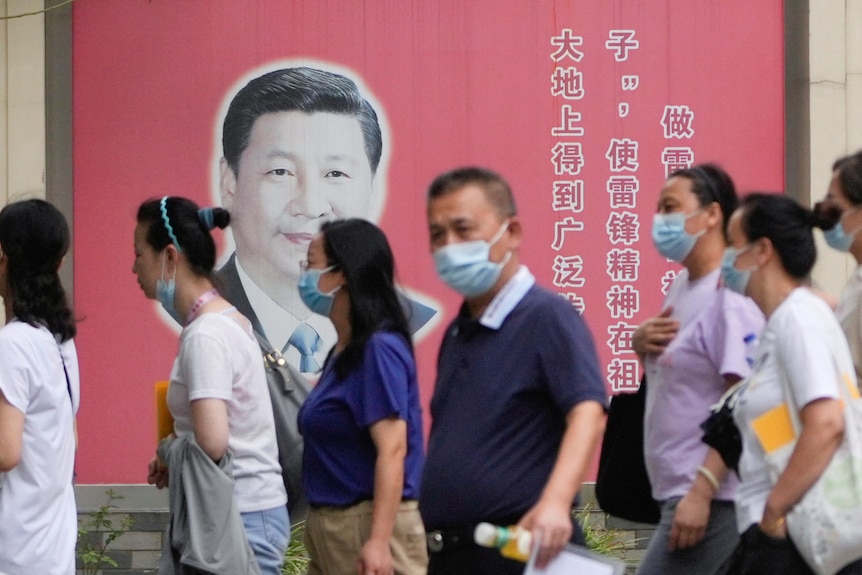 A crowd of Chinese people wearing masks pass a picture of Xi Jinping on a red poster.