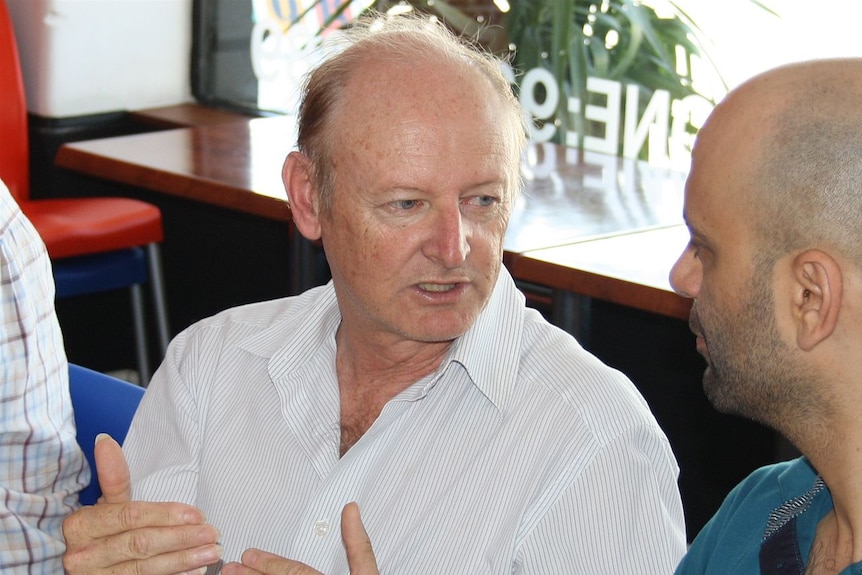 A man in a white shirt gestures with his hands as he chats with another man.