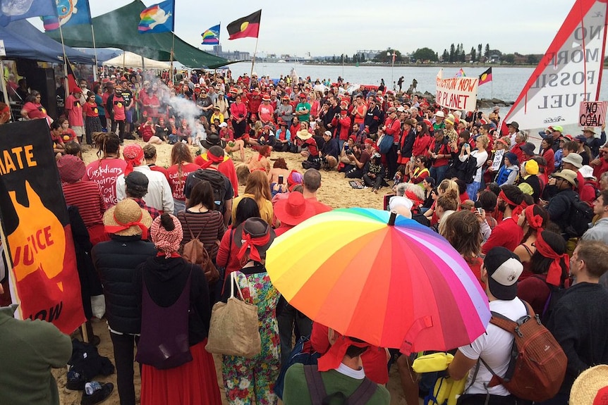 Hundreds of people gather around a fire by the beach.