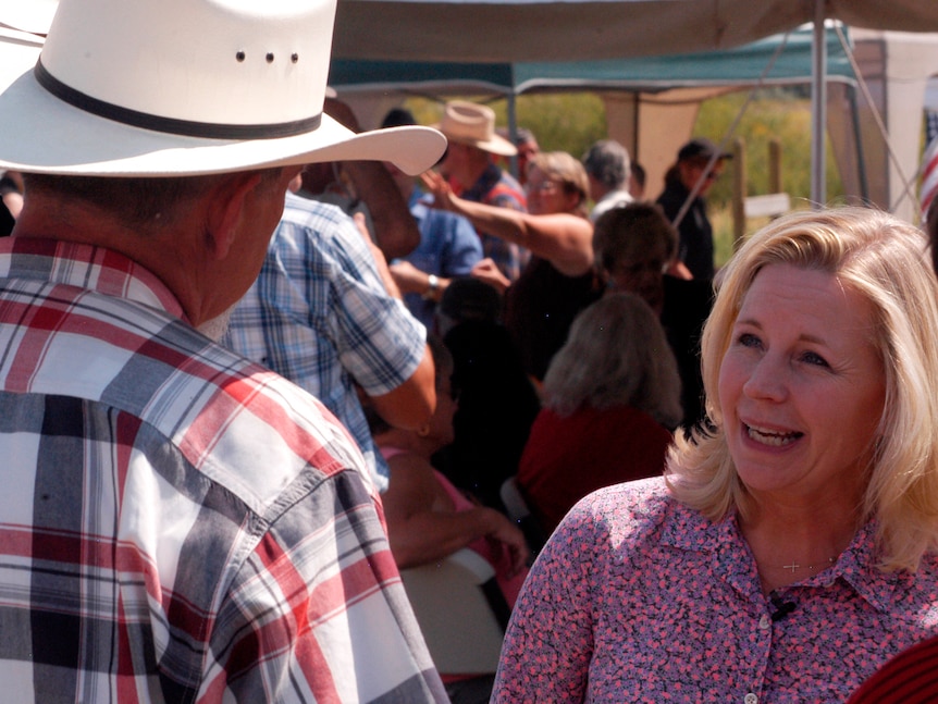 Liz Cheney, in a floral shirt, speaks to a man wearing a wide-brimmed hat and a checked shirt