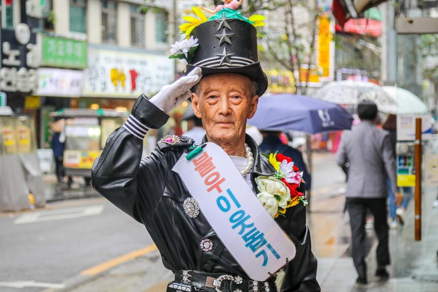 An older man in a top hat, white gloves, sash and flower arrangement salutes in front of the camera