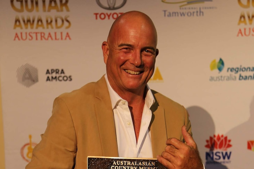 James Blundell wears a tan blazer and white shirt as he smiles while holding Australian Country Music Roll of Renown award.
