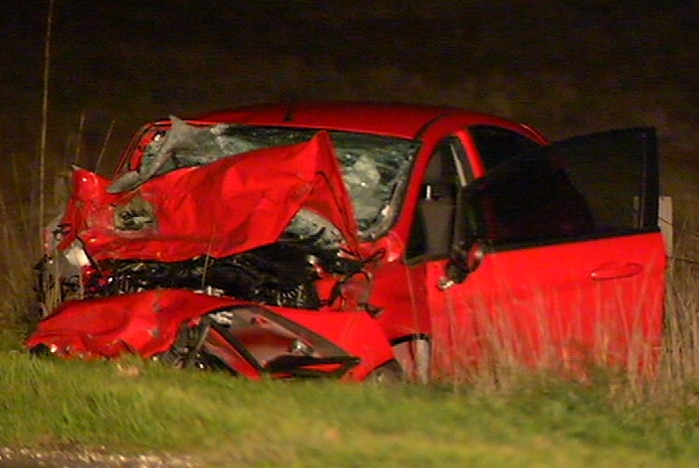The wreckage of a small red car by the side of the road at the scene of the collision.