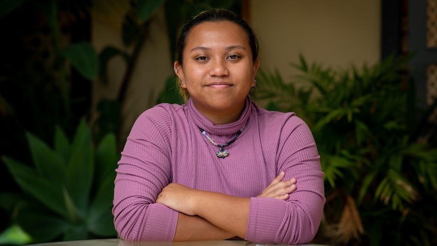 A woman sits at a table with her arms folded staring straight into the camera, with green plants in the background