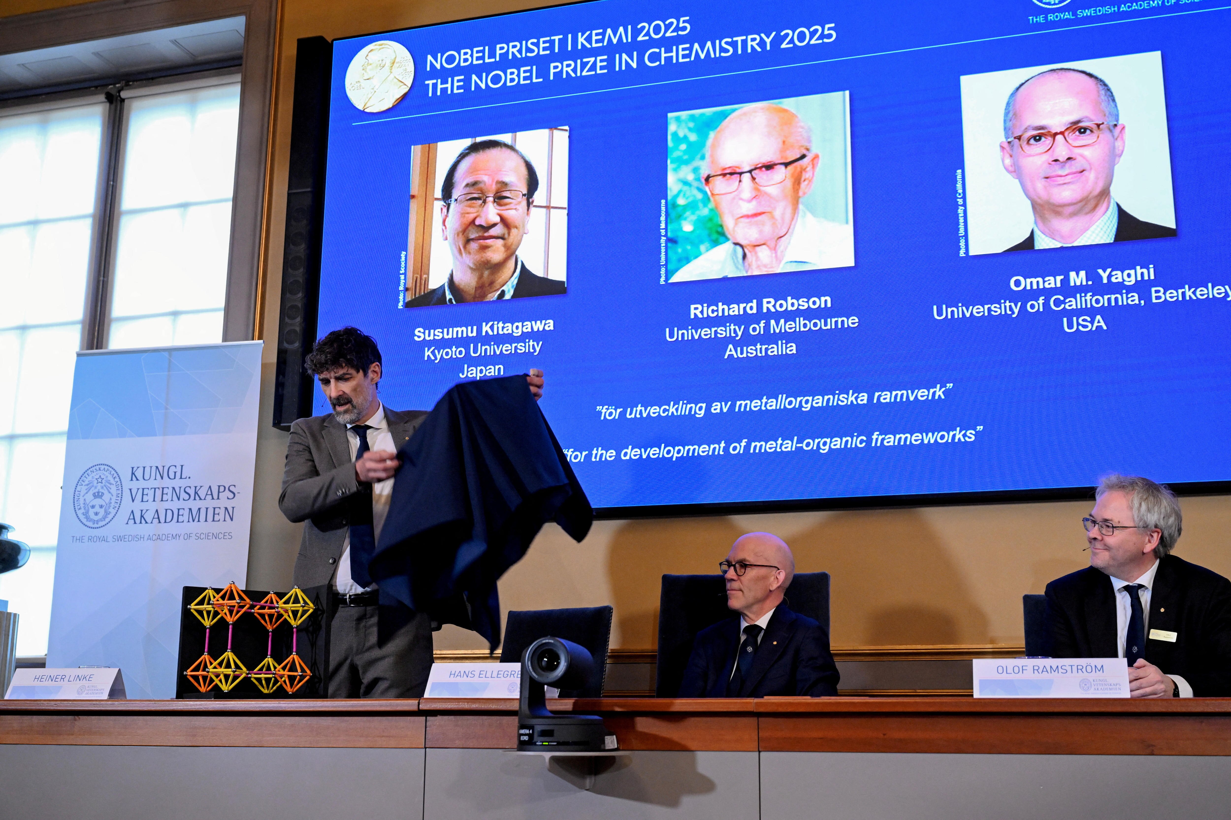 A man lifts a cloth off a diorama of cells as a screen behind him shows three Nobel Prize laureates