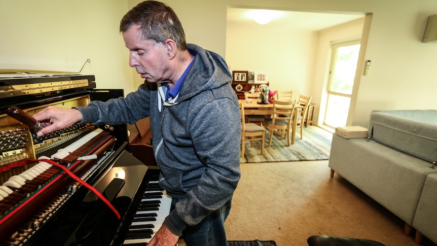 Graeme McGowan bending over an opened piano with his right hand on the tuning hammer and his left hand on the keys.