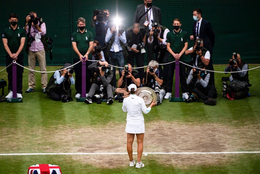 Ash Barty is seen from behind holding the round Wimbledon plate in front of a bank of photographers