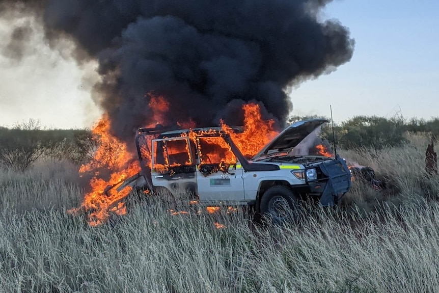 a white car is engulfed by flames in a grassy desert area.