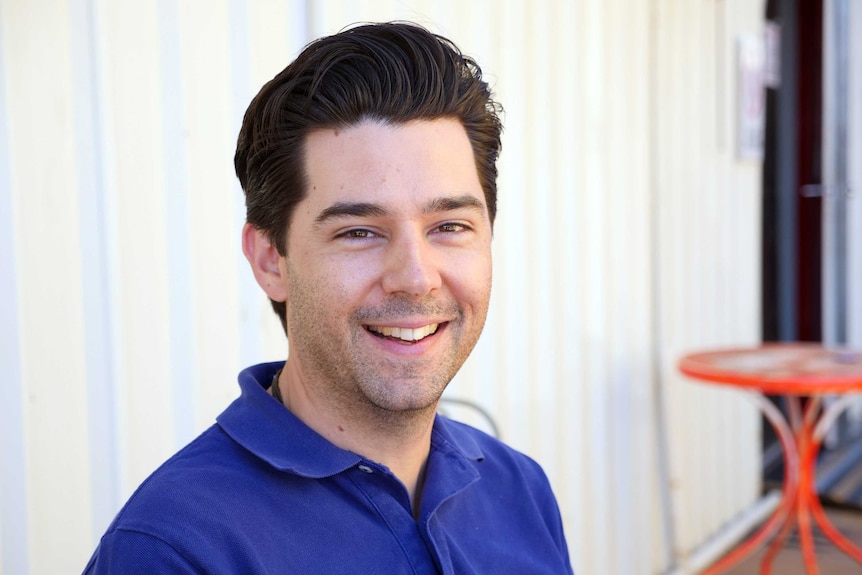 A man with dark eyes and hair smiles directly at the camera wearing a blue polo shirt.