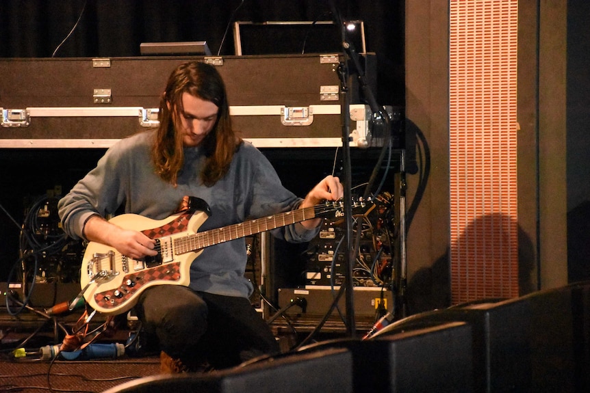 A musician with long hair kneels down and tunes his guitar onstage.