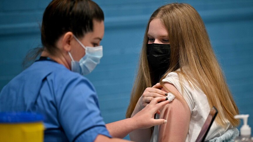 A young woman with long blonde hair is given a COVID vaccine by a nurse dressed in blue scrubs