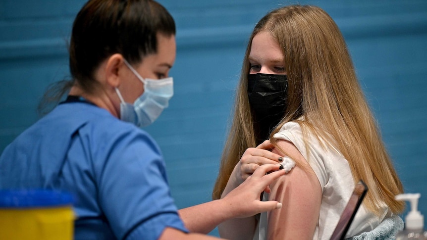 A young woman with long blonde hair is given a COVID vaccine by a nurse dressed in blue scrubs