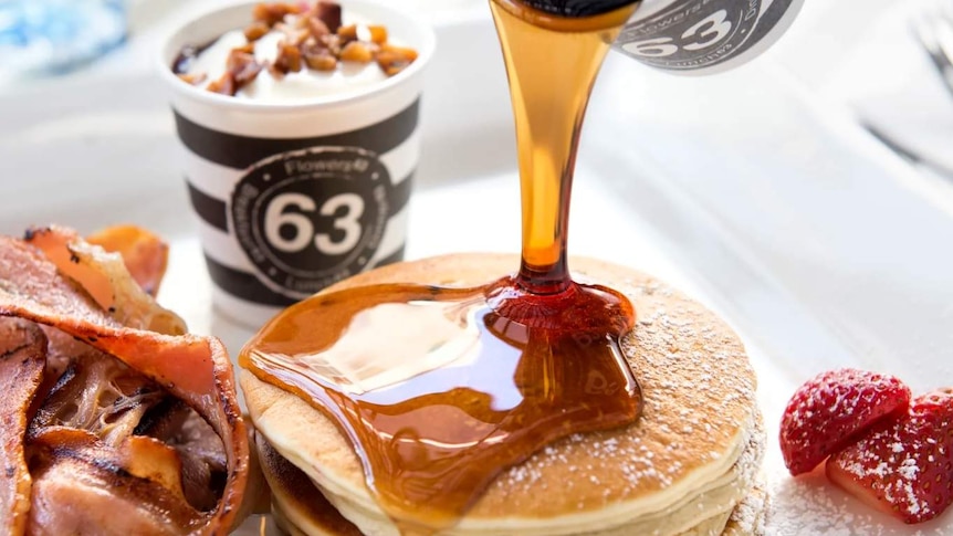 Close-up of maple syrup being poured onto pancakes, with a cup behind it carrying the Cafe 63 logo.