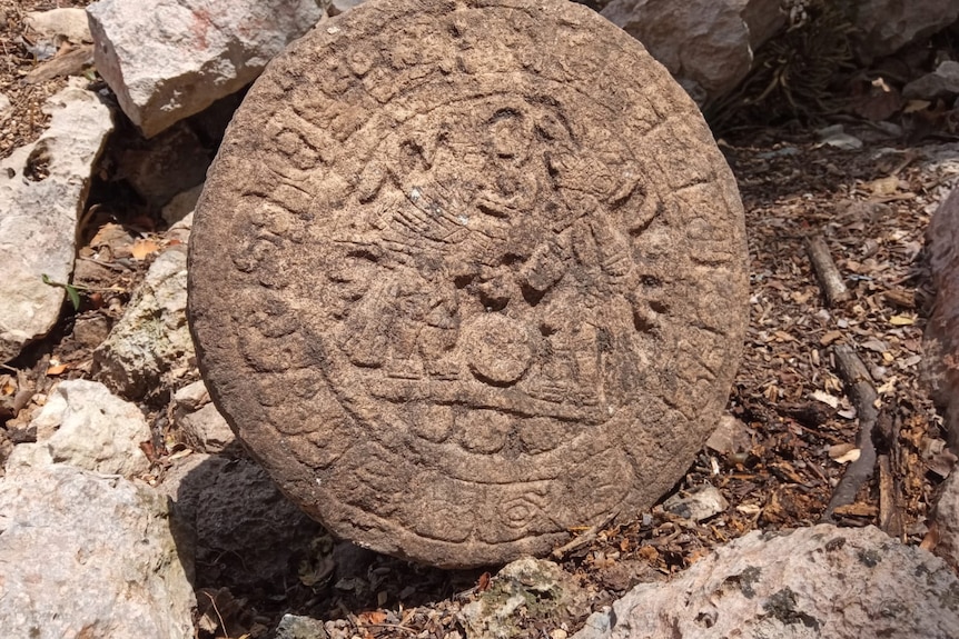A round stone tablet with carved designs tests on a piece of rocky ground.