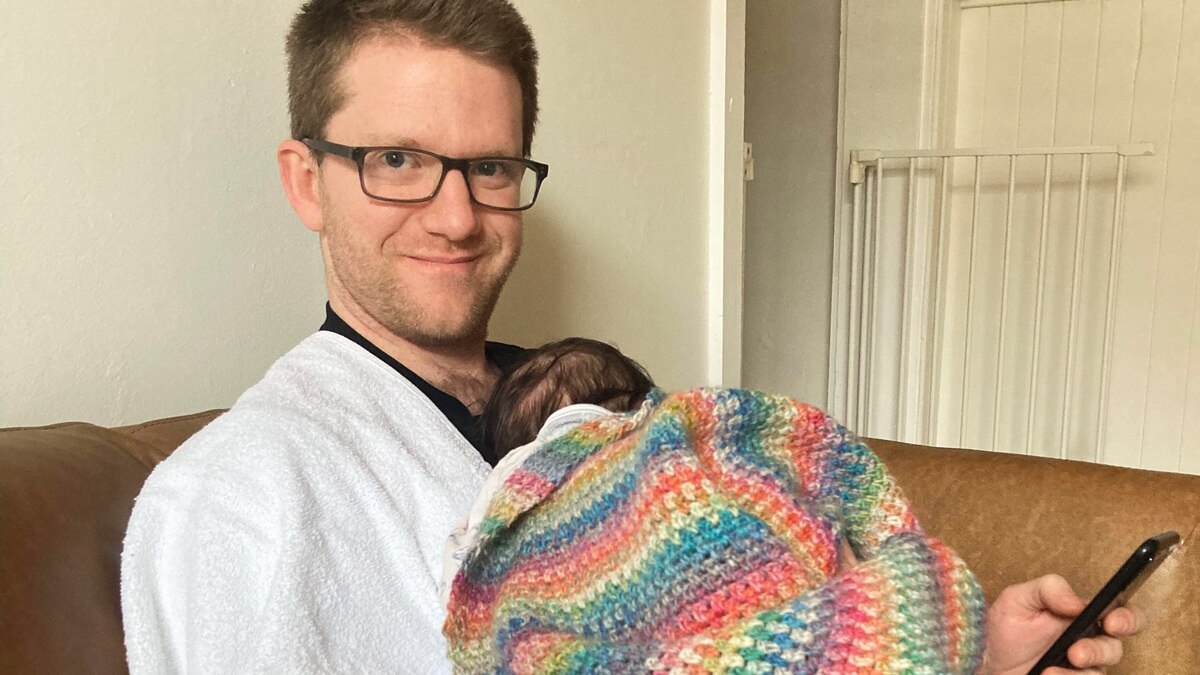 Alex McKinnon sits on a couch holding his newborn daughter, wrapped in a crocheted blanket with her head against his chest.
