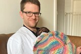 Alex McKinnon sits on a couch holding his newborn daughter, wrapped in a crocheted blanket with her head against his chest.