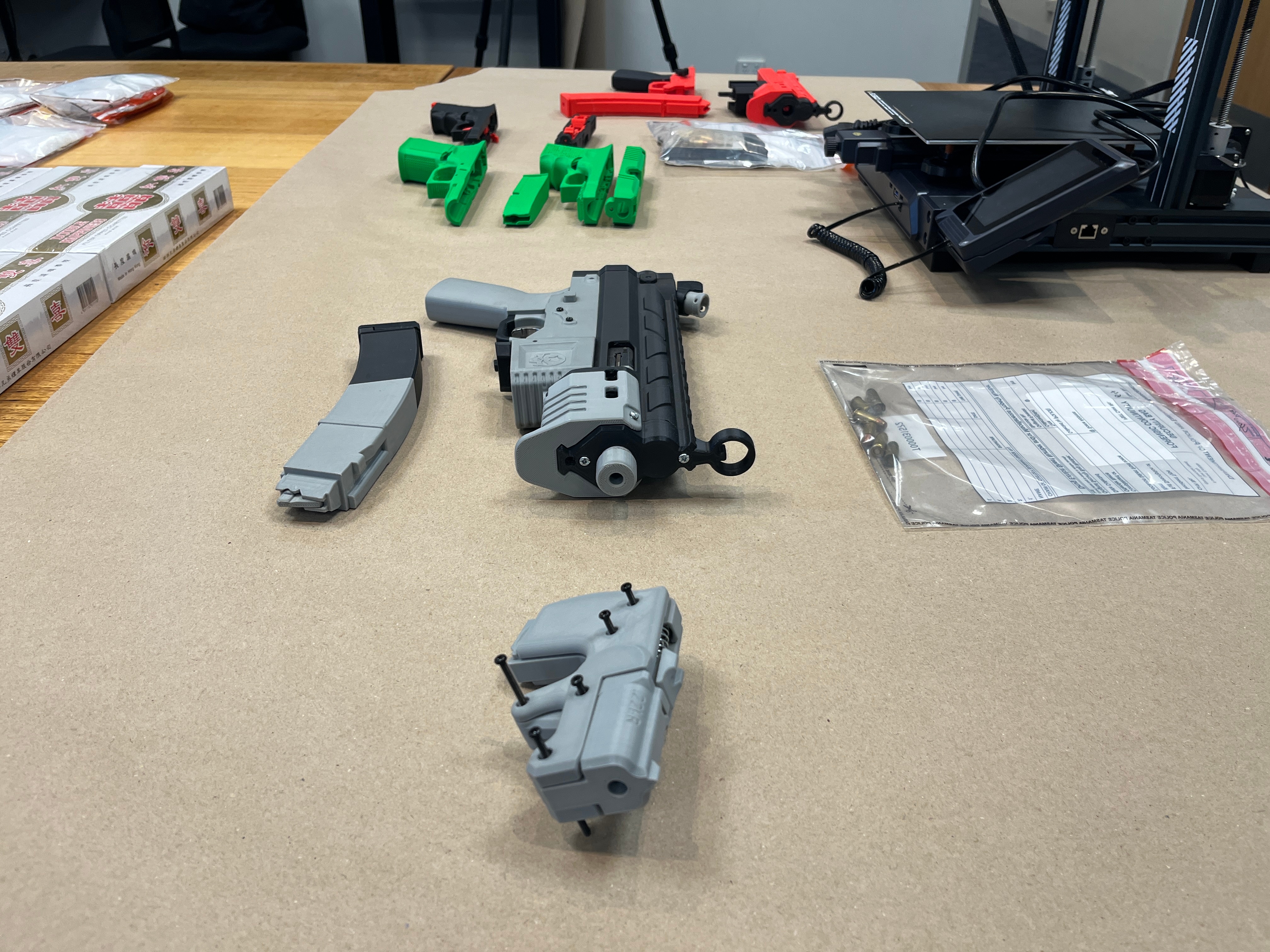 A dozen 3D printed firearms and a 3D printer laid out on a wooden table.
