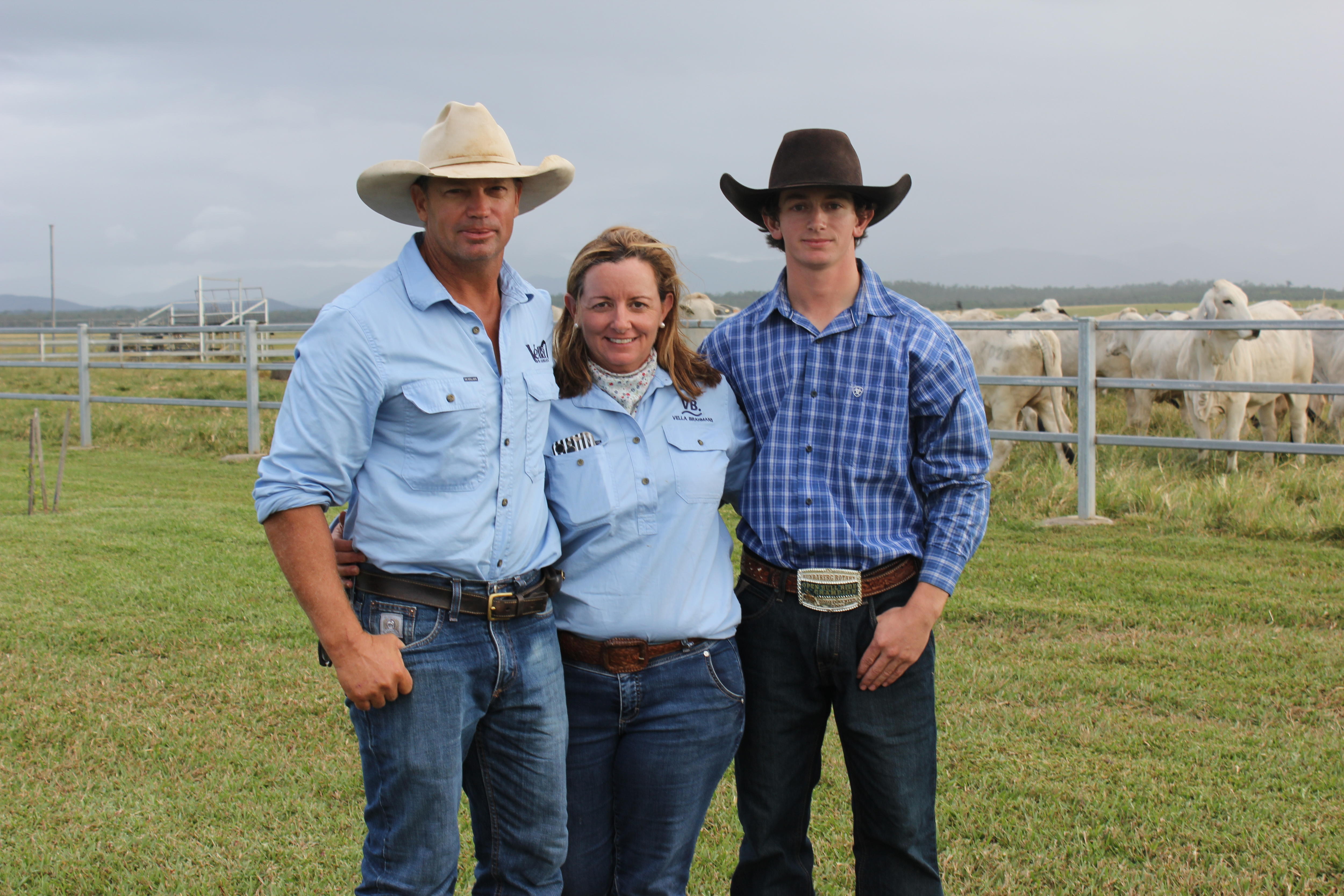 A husband, wife and teenage son with farm shirts, jeans and hats on in front of a paddock of white cattle.