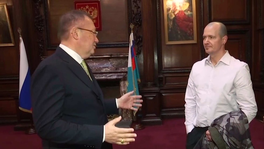 Charlie Rowley, standing to the left, wears a white shirt and looks at Alexander Yakovenko, inside an elaborate office