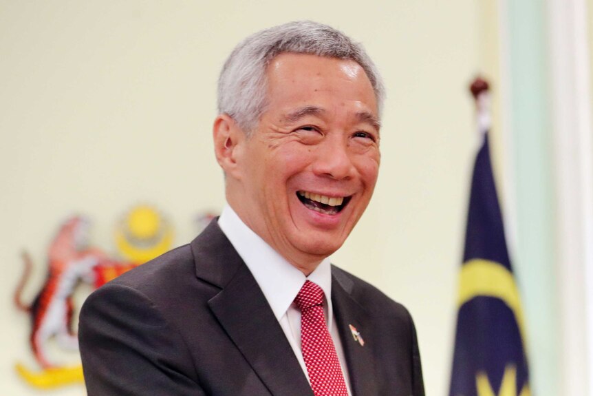 Singapore's PM Lee Hsien Loong laughs in front of a Malaysian flag and crest of arms.