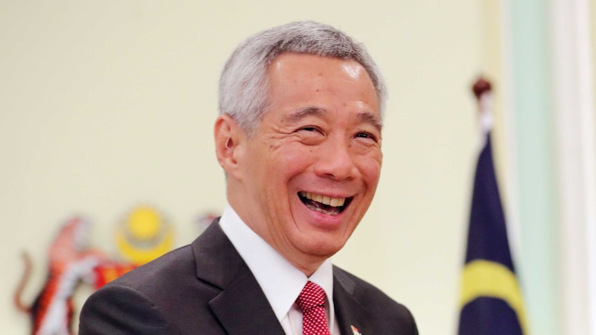 Singapore's PM Lee Hsien Loong laughs in front of a Malaysian flag and crest of arms.