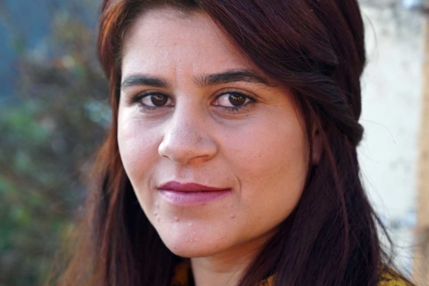 Portrait of Yazidi woman Hayfa Adi in 3/4 profile looking at camera with a wall and plants in the background