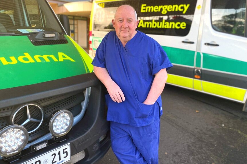 A man in hospital scrubs leans against a parked ambulance with his hand in his pocket.
