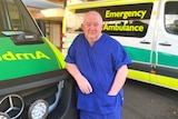 A man in hospital scrubs leans against a parked ambulance with his hand in his pocket.