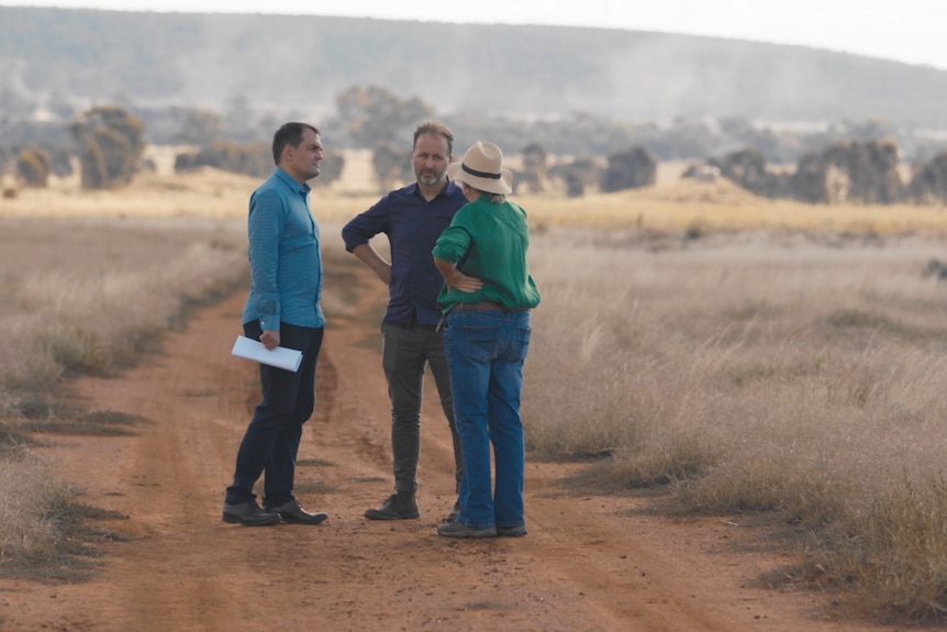 Two men and a woman stand on a dirt road, with a hill in the background