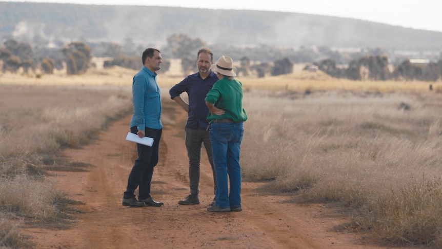 Two men and a woman stand on a dirt road, with a hill in the background