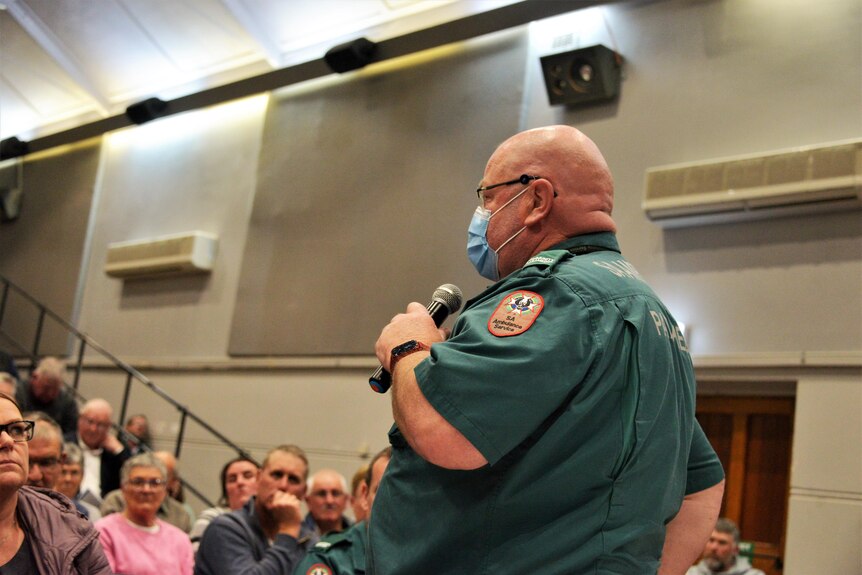 A man in an ambulance uniform holds a microphone addressing an audience 