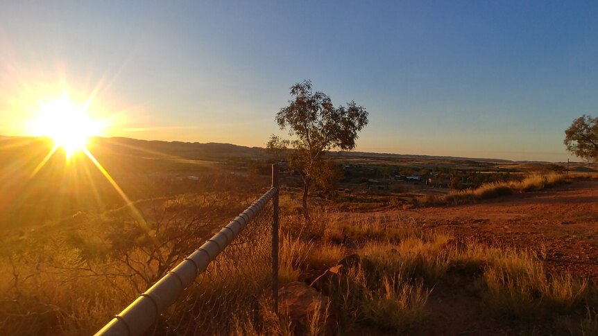 The sun sets on the left, with a fence line, tree and dirt track in front
