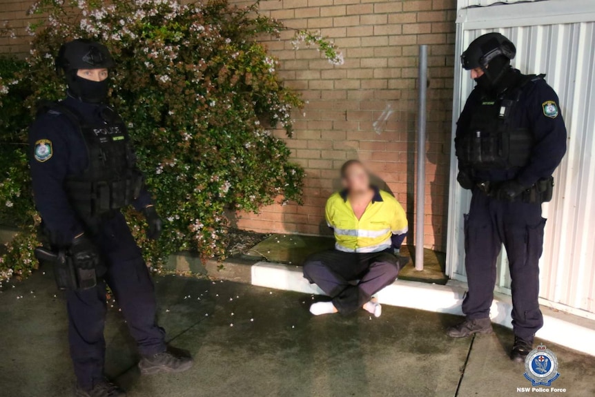 Two people in black police outfits standing on either side of one person sitting on the ground.