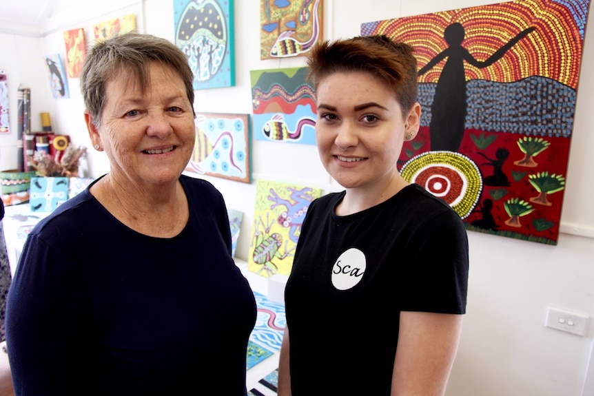 A mother and daughter, Kathy Pemberton and Jamira Pemberton, standing together in front of Aboriginal style paintings.