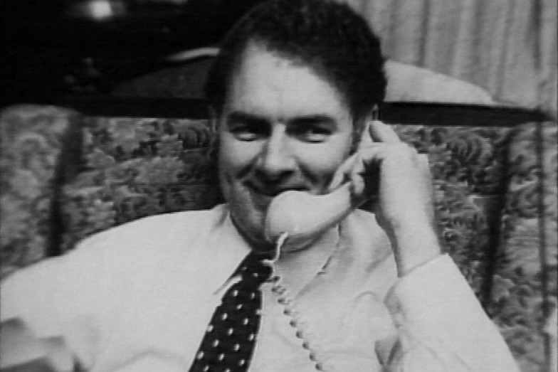 A man with dark hair wearing a shirt and tie is talking on the phone while smiling.