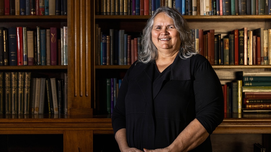 An Indigenous woman with shoulder-length grey hair poses for a portrait in front of shelves of books, smiling warmly.