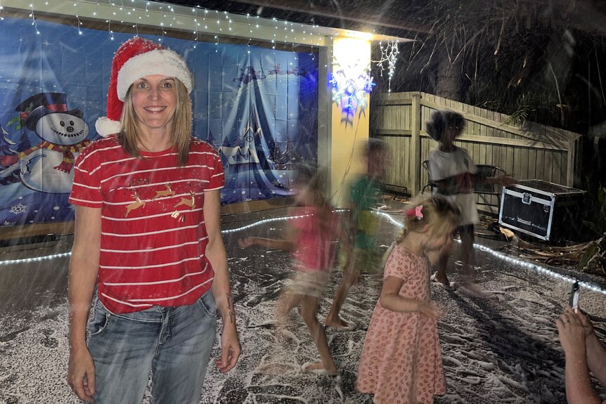 Widely smiling woman, blonde hair, striped red and white t-shirt, jeans, santa hat in front of playing children.