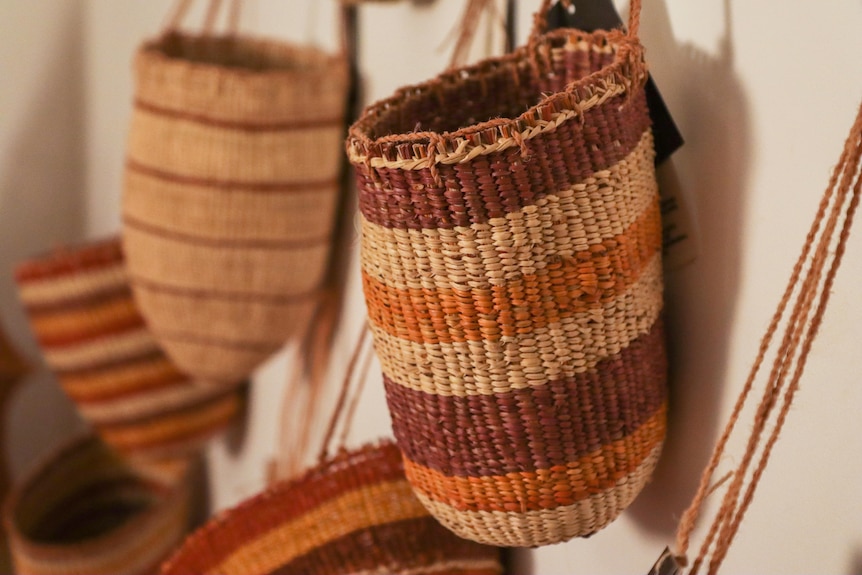 A variety of hand-woven dilly bags with brown, yellow and wheat-coloured thread are hung on the wall of a gallery.