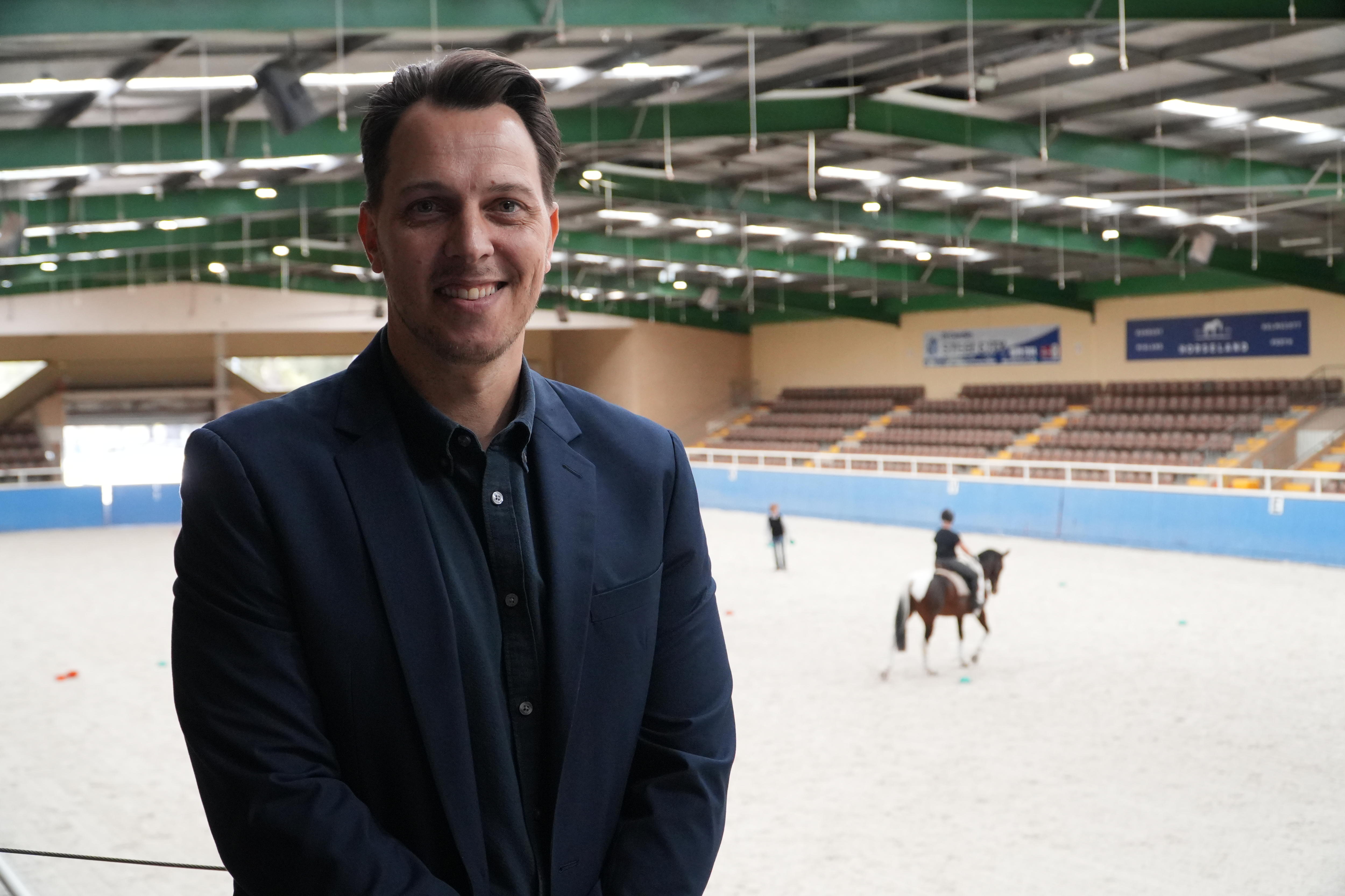 A man smiles to camera in an equestrian facility.