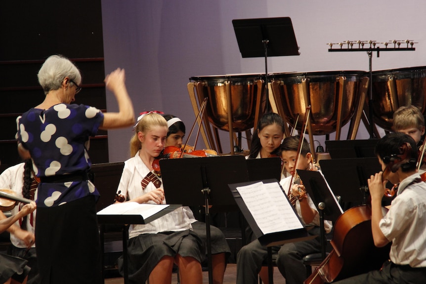 Young students performing in a school orchestra with a conductor in front of them.