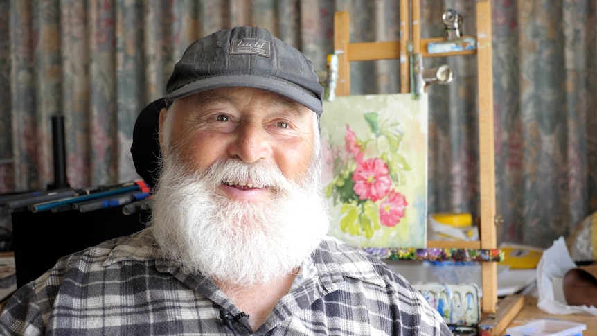 A 67-year-old man with a white beard and gentle smile sits in front of his painting of pink flowers